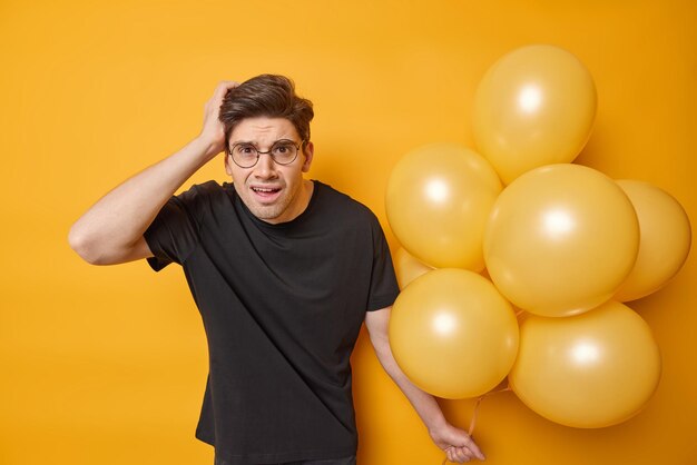 Indignant displeased European man keeps hand on head reacts on unpleasant news wears black casual t shirt holds bunch of inflated balloons isolated over yellow background prepares for party.