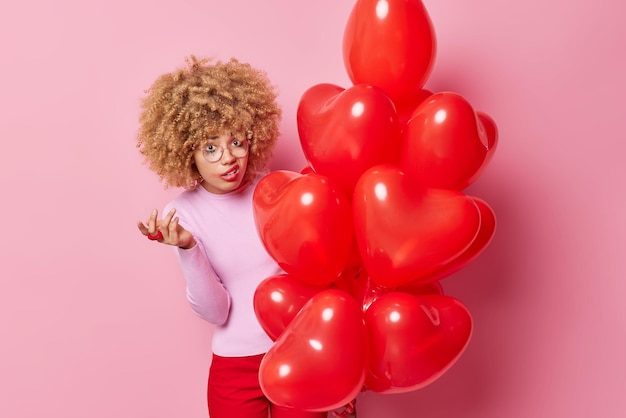 Free photo indignant frustrated curly woman shrugs shoulders looks puzzled at camera holds big bunch of heart balloons doesnt like something purses lips has leaked makeup isolated over pink background