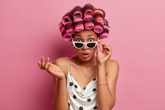 Indignant puzzled woman stares with shock, keeps hand on sunglasses, cannot believe her eyes, wears stylish sunglasses, makes hairstyle with hair rollers, wears casual dress, stands indoor