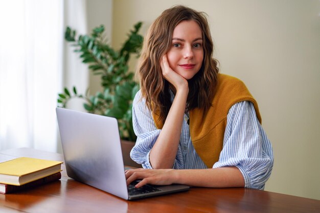 Indoor home office portrait of trendy blogge influencer working on her laptop, stylish clothes, minimalistic interior.