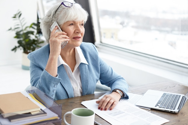 Indoor image of gray haired sixty year old mature female banker working in stylish office, discussing details of civil case with her client on mobile phone, sitting at desk by window, using laptop