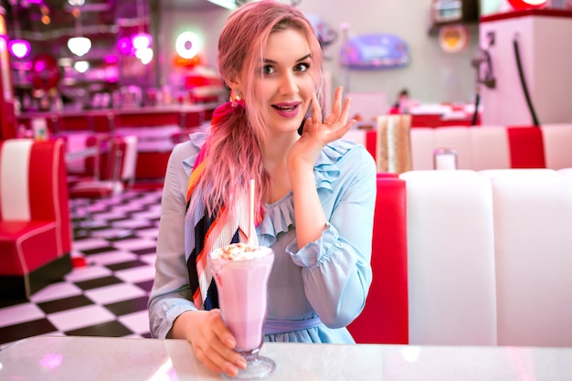 Free photo indoor image of pretty young elegant woman enjoying her tasty sweet strawberry milk shake at retro vintage american restaurant, neon design, cute pastel dress, pink hairs and accessories