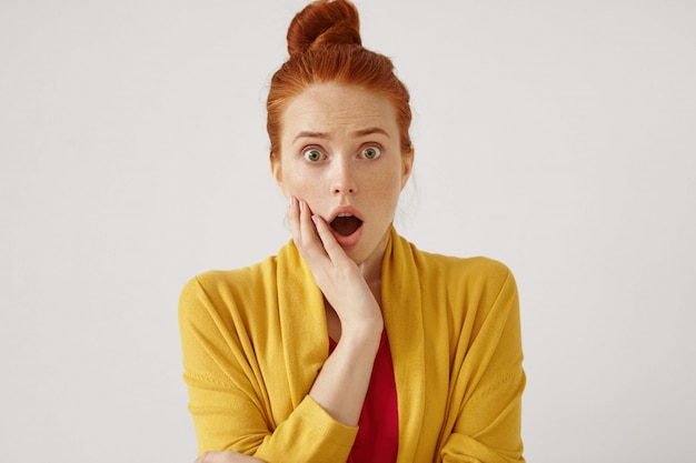 Free photo indoor isolated shot of attractive shocked young redhead woman with hair bun opening mouth wide opened and holding hand on her cheek, frozen by fear, shock or astonishment. amazement concept