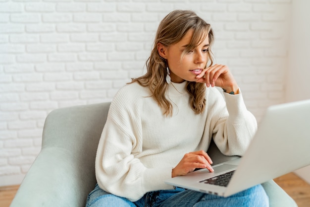 Free photo indoor lifestyle photo of pretty woman with blond wavy hairs sitting on sofa at home working on laptop computer