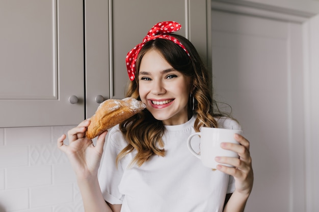 Indoor photo of wonderful girl eating sweet croissant. Portrait of amazing european lady having fun during lunch.