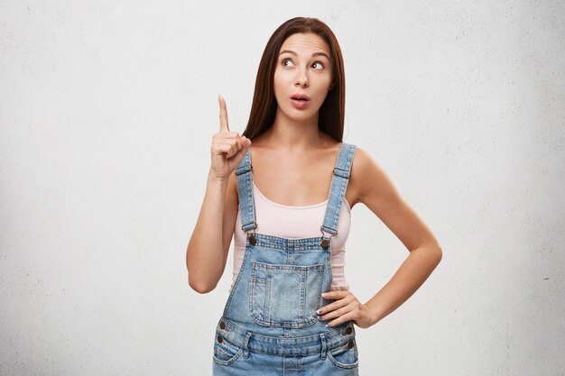 Indoor picture of attractive young brunette woman of European appearance looking away and raising index finger, having bright idea or interesting thought, standing isolated at white wall