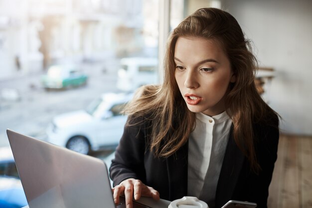 Indoor portrait of bothered and confused stylish woman sitting in cafe, working with laptop, looking at screen with surprised expression