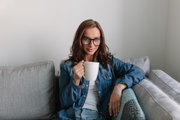 Indoor portrait of caucasian girl with snowwhite smile looks into camera Against white background at home darkhaired beauty in round glasses white tshirt and blouse