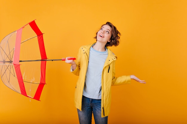 Free photo indoor portrait of charming girl looking up during rainy weather.  caucasian pale woman in yellow jacket posing with parasol and happy face expression.