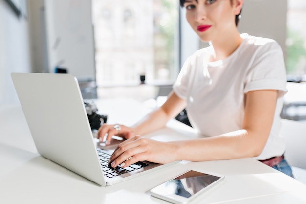 Indoor portrait of elegant brunette woman in white shirt with modern laptop and smartphone on foreground