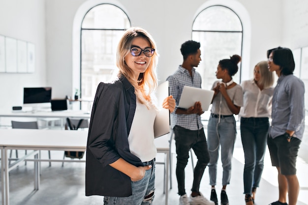 Free photo indoor portrait of elegant business-lady in black jacket and her team. african office manager in white sneakers carrying laptop and talking with mulatto woman in jeans.