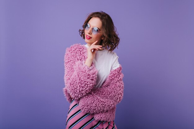 Indoor portrait of elegant caucasian woman wears trendy bright jacket Dreamy girl in sunglasses posing with pleasure in fur coat on purple background