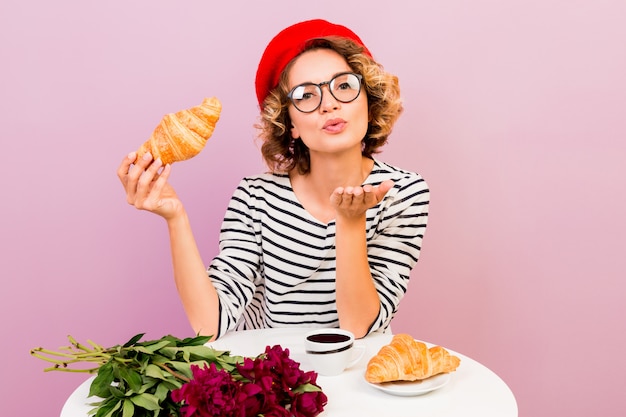 Free photo indoor portrait of elegant french woman in red beret woman in spectacles blows sweet kiss and holding croissants.