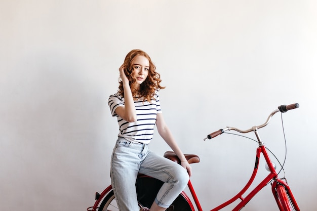 Free photo indoor portrait of elegant lady in jeans sitting on bicycle. adorable ginger woman posing with bike.