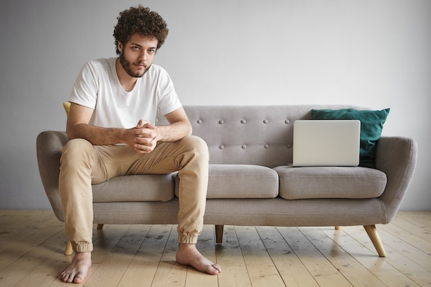 Free photo indoor portrait of fashionable self employed young bearded man in casual clothes working from home, using wifi on portable electronic device, sitting on sofa in living room with generic laptop