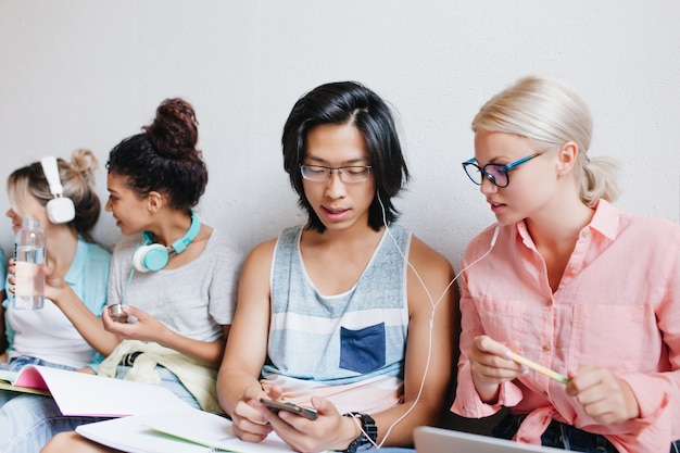 Indoor portrait of group of people with blonde woman and asian boy. Attractive fair-haired female student in elegant glasses enjoying music in earphones with university friend.