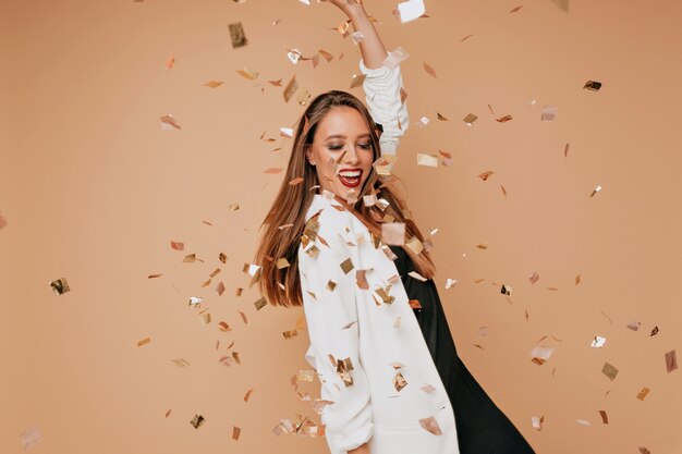 Indoor portrait of pretty young female model with light-brown hair wearing white jacket and black dress dancing and having fun on beige wall with confetti