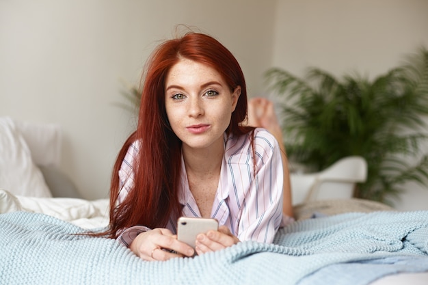 Indoor portrait of relaxed beautiful young redhead European female in nightwear ordering food delivery online, using application on her mobile phone, lying on undone bed late in the morning