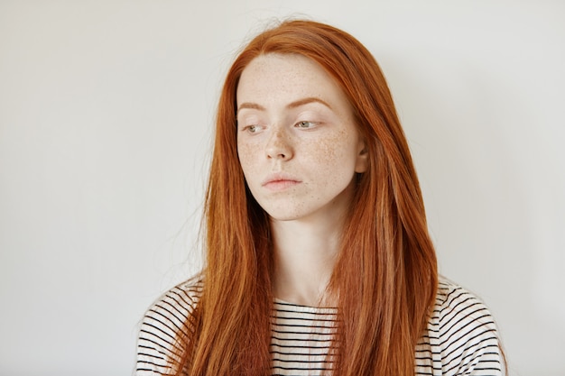 Indoor portrait of sad young woman wearing her long ginger hair loose looking down with unhappy face expression