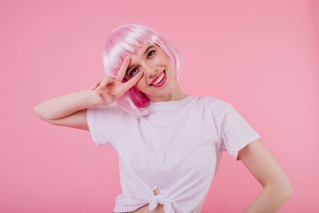 Indoor portrait of smiling lovely girl with pink hair isolated on pastel wall. graceful caucasian lady in white t-shirt posing with peace sign and laughing