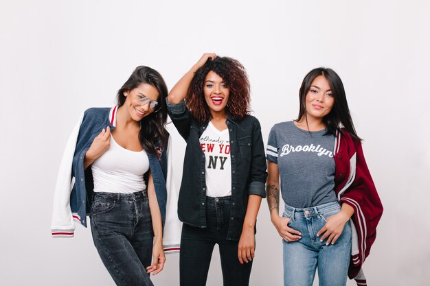 Indoor portrait of three excited female students in trendy clothes having fun together after lessons. Curly girl in denim attire spending time with brunette friends and laughing.