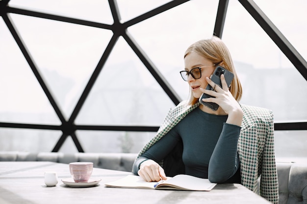 Indoor portrait of a young businesswoman sitting in a cafe and talking on a phone. Blonde girl wearing sunglasses and jacket