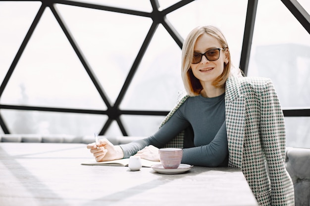 Free Photo indoor portrait of a young businesswoman sitting in a cafe and writing a documents. blonde girl wearing sunglasses and jacket