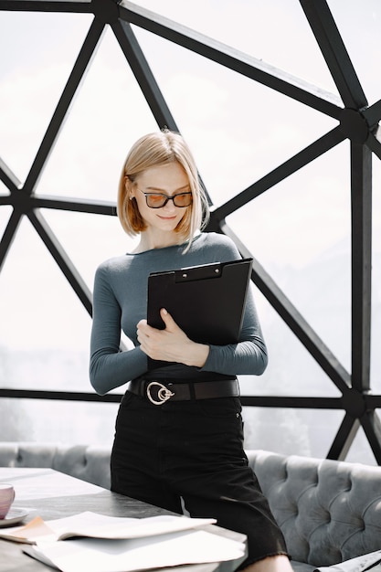 Indoor portrait of a young businesswoman standing in a cafe and writing on a paper holder. Blonde girl wearing sunglasses