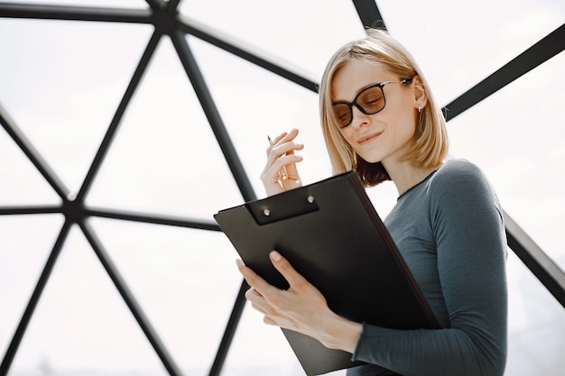 Indoor portrait of a young businesswoman standing in a cafe and writing on a paper holder. Blonde girl wearing sunglasses