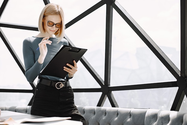 Indoor portrait of a young businesswoman standing in a cafe and writing on a paper holder. Blonde girl wearing sunglasses