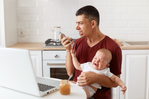 Indoor shot of aggressive sorrow brunette male wearing maroon casual style t shirt sitting at table in kitchen with his infant daughter, screaming while recording voice message.