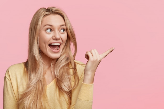 Indoor shot of amazed blonde woman has overjoyed expression, indicates with excited look aside, poses against pink wall with blank copy space for your advertising content. Wow, that`s great!