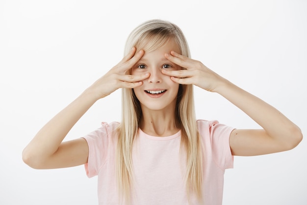 Free Photo indoor shot of amazed surprised cute daughter with broad happy smile, holding fingers near eyes and grinning from positive scene, being joyful, attending great party in her honor over gray wall