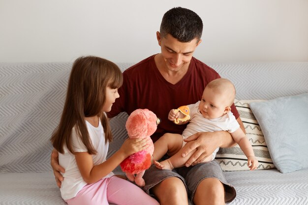 Indoor shot of attractive brunette man wearing maroon t shirt spending time with his children, sitting on sofa in light room, playing together, expressing happiness.