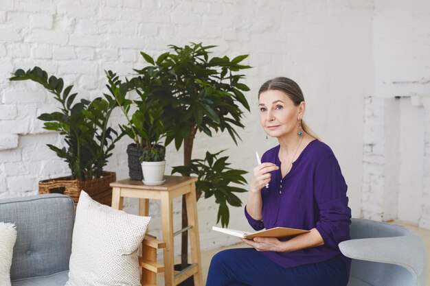 Indoor shot of attractive pensive middle aged European female business coach sitting in light spacious room, surrounded with modern furniture and plant pots, checking schedule in her diary