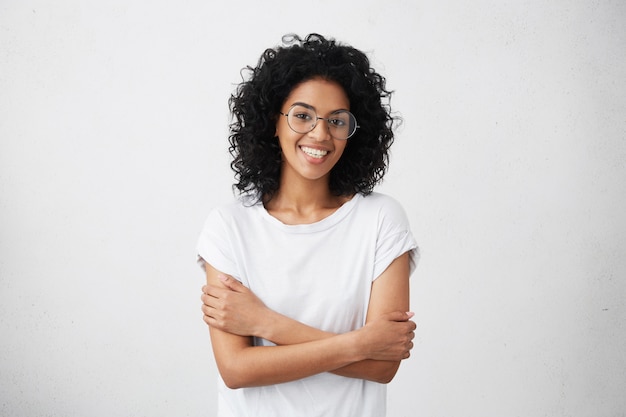 Indoor shot of beautiful happy African American woman smiling cheerfully, keeping her arms folded, relaxing indoors after morning lectures at university