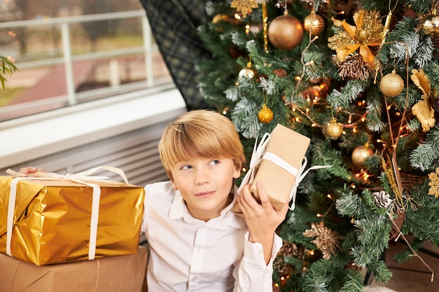 Indoor shot of blonde teen boy sitting under decorated New Yearâs tree surrounded with Christmas gifts, shaking box, trying to guess what is inside, having curious interested facial expression