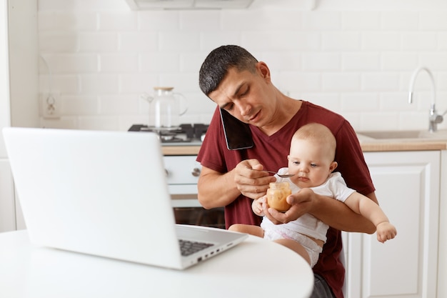 Indoor shot of brunette male wearing maroon casual style t shirt sitting at table in kitchen, talking via smartphone phone feeding his infant daughter with fruit puree.