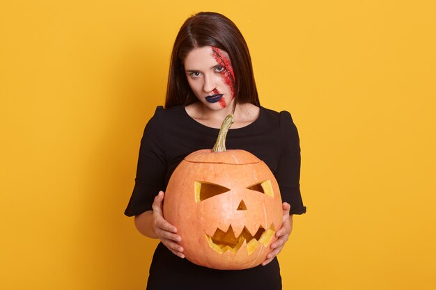 Indoor shot of brunette woman in Halloween makeup holding pumpkin over yellow