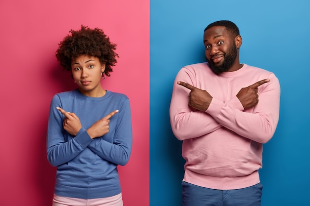 Free photo indoor shot of confused ethnic couple cross hands over chest, point left and right, show directions, cannot make choice