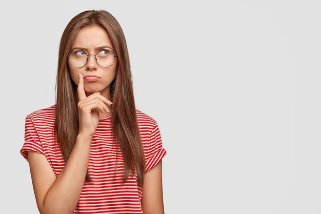 Indoor shot of contemplative pleasant looking woman keeps index finger near mouth