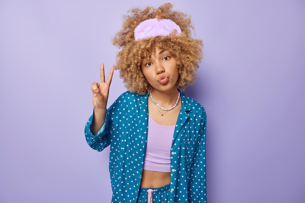 Indoor shot of curly haired woman wears sleepmask and comfortable pajama keeps lips folded gestures victory or peace sign isolated over purple background Good morning and awakening concept