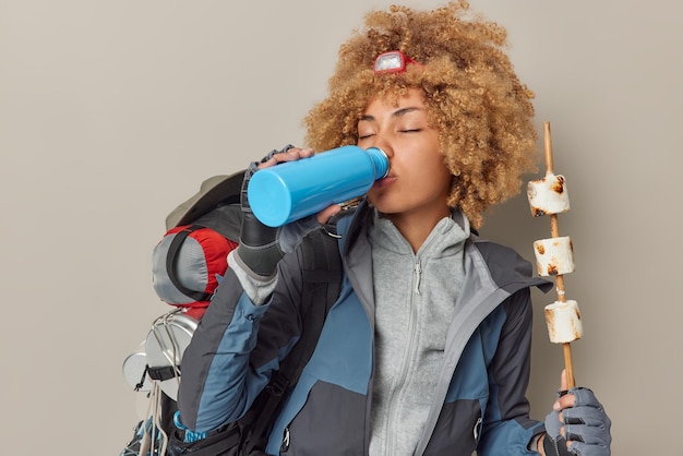 Free Photo indoor shot of curly woman camper drinks water from bottle holds roasted marshmallow on stick has picnic time wears jacket and gloves poses with backpack people hiking and summer rest concept