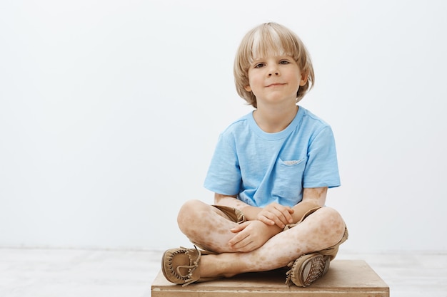 Indoor shot of cute happy blond child with positive smile sitting with crossed hands, having vitiligo, smiling broadly while hanging out with pals in kindergarten