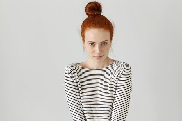 Indoor shot of cute redhead girl with hair knot wearing casual striped long-sleeved t-shirt, her posture expressing shyness. Beautiful young woman posing at blank wall