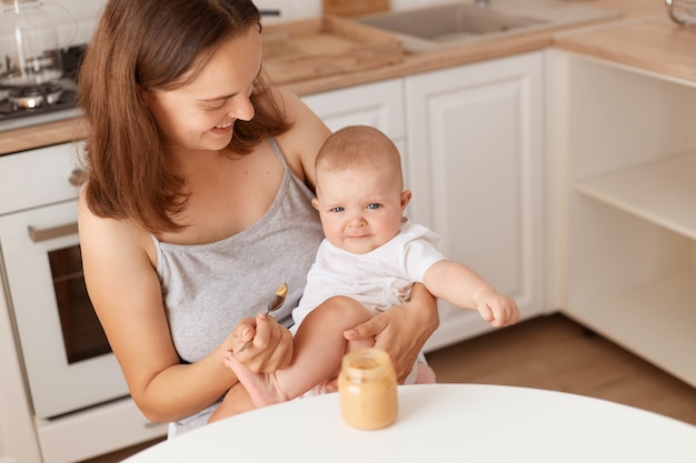 Free photo indoor shot of dark haired female feeds her little daughter with fruit or vegetable puree, mother looking at her cute baby with love, healthy feeding up.