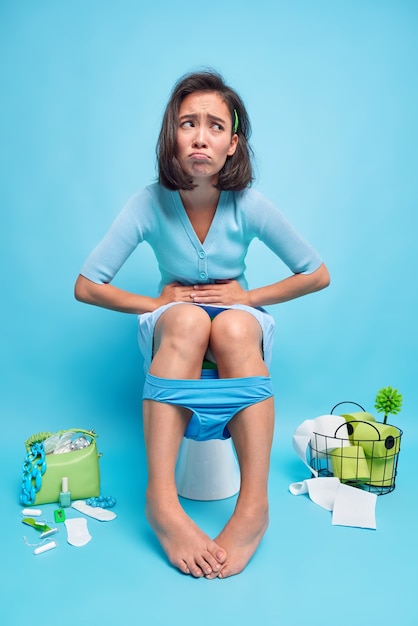 Indoor shot of displeased Asian woman suffers from stomachache indigestion or diarrhea poses poses toilet bowl feels unwell because of abdominal pain wears panties down on legs