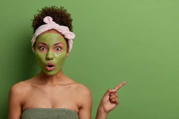 Indoor shot of emotional surprised woman does beauty procedures, applies facial mask for rejuvenation, demonstrates something shocking on empty space, green wall. Skin care, wellness concept