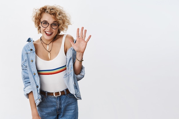 Free photo indoor shot of enthusiastic stylish female blonde with glasses and pierced nose raising palm and smiling broadly giving recommendation and positive opinion about promotion