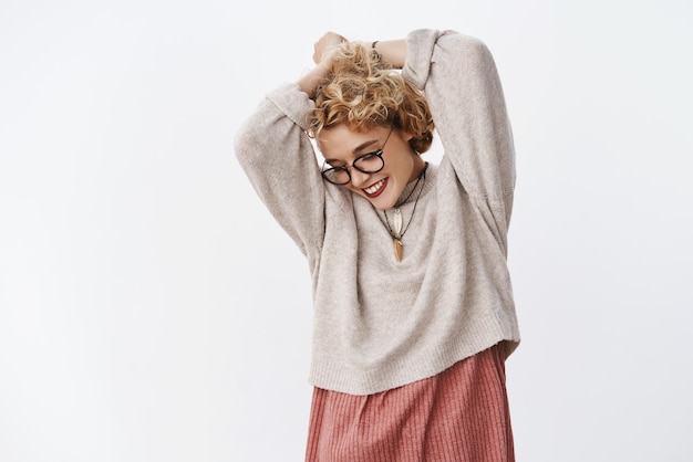 Free Photo indoor shot of excited enthusiastic and carefree charming tender woman with short blond hairstyle in hipster glasses and sweater raising hands behind head looking down with pretty sensual smile